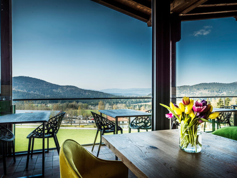 Ein Tisch mit Blumen vor einem großen Fenster mit Panoramablick auf den Bayerischen Wald im natura Hotel Bodenmais.